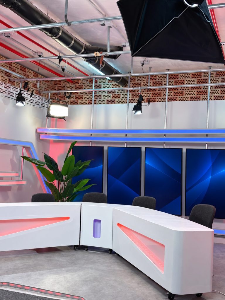 Detalle del plató: mesa blanca con luces LED rosas, sillas, planta decorativa y cuatro pantallas verticales azules bajo la parrilla de iluminación del techo.