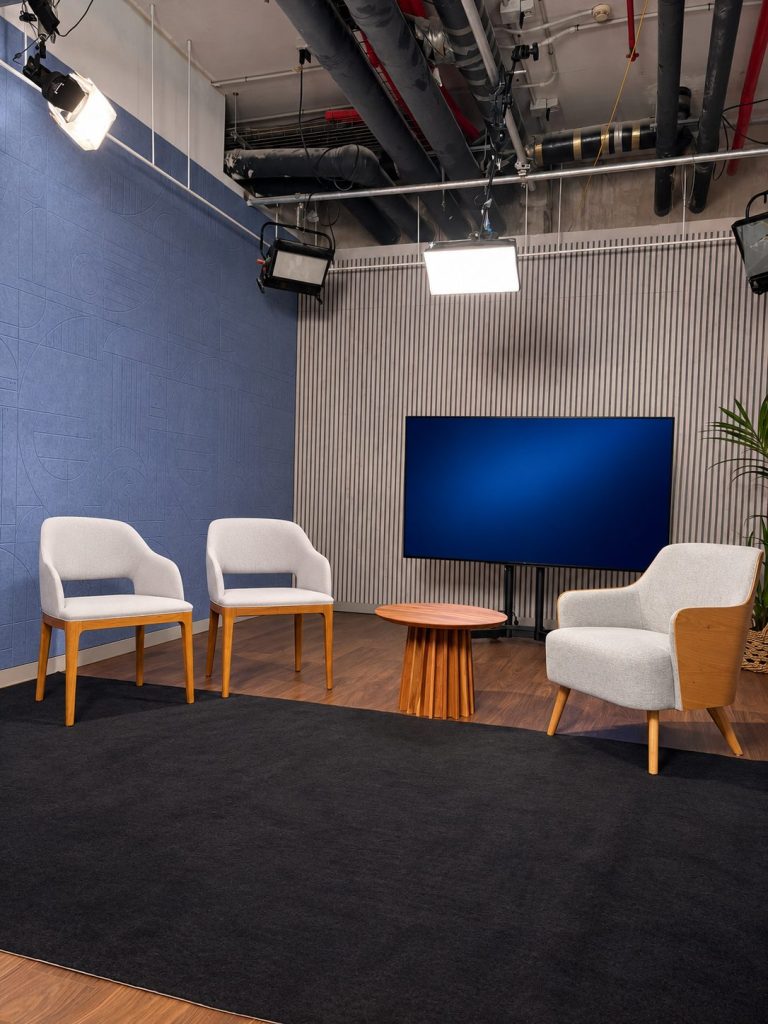 Set de entrevistas con tres butacas tapizadas en gris claro, mesa auxiliar redonda de madera y pantalla grande al fondo, entre una pared de paneles acústicos azules y otra de listones grises, con focos de estudio colgados del techo.
