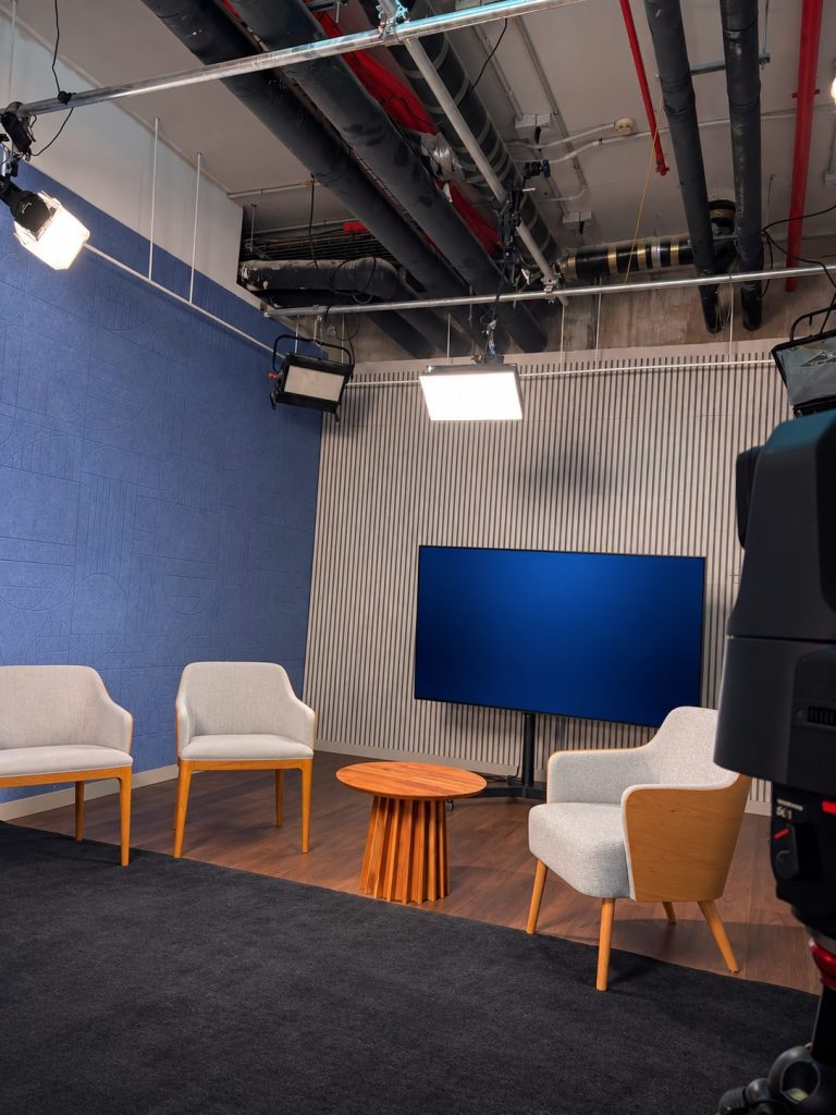 Set de entrevistas visto desde detrás de una cámara de estudio: tres butacas, mesa auxiliar de madera y pantalla al fondo, iluminado por focos LED suspendidos del techo.